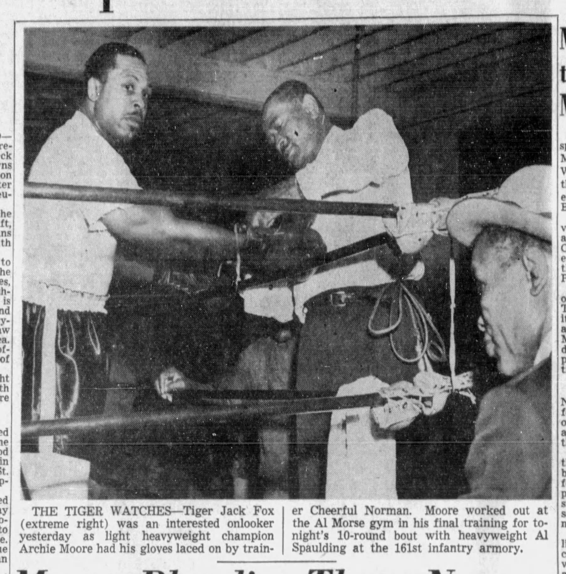 Two Black men are standing in a boxing ring, one lacing boxing gloves onto the other's hands. Tiger Jack Fox is seated just outside the ring, watching intently.