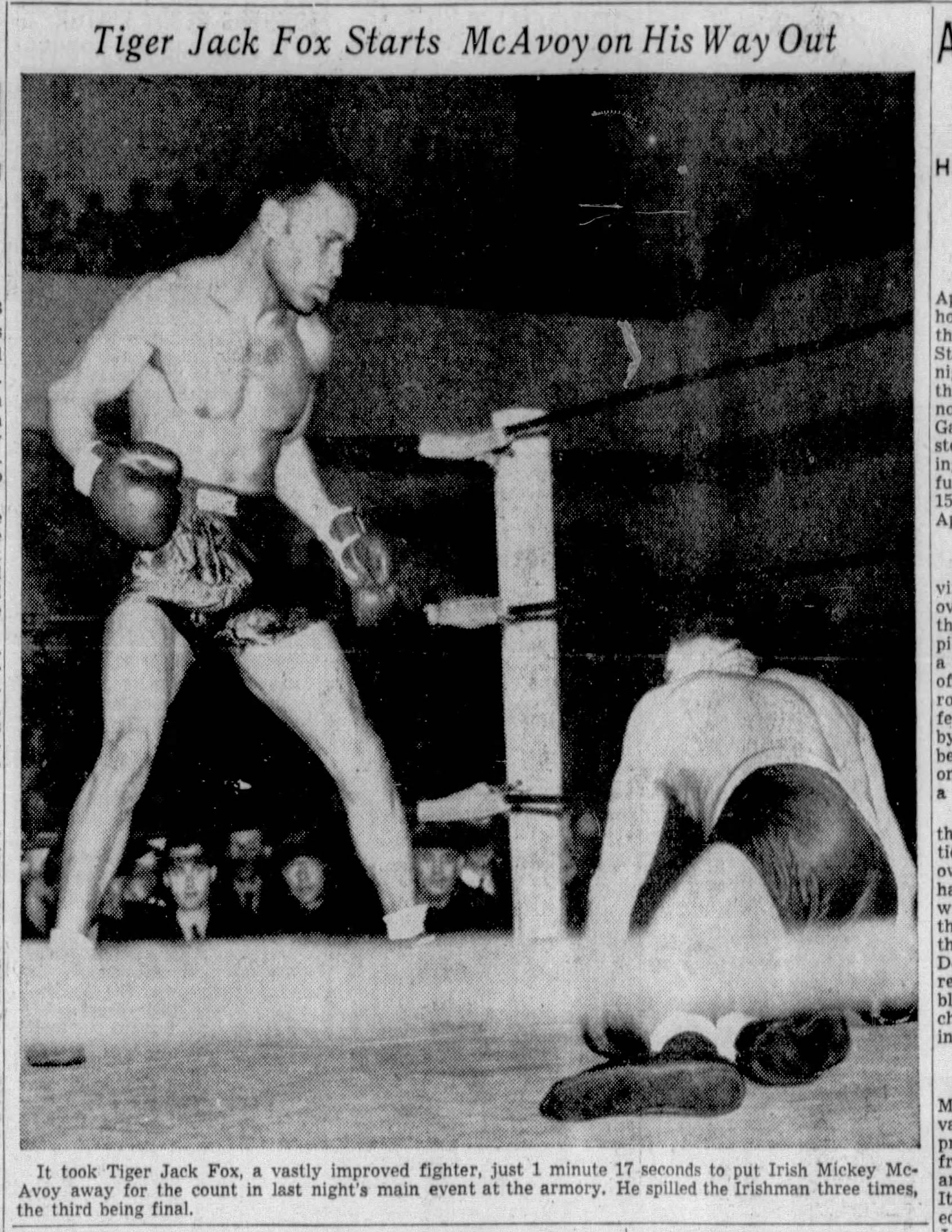 A large Black man in boxing shorts and gloves is standing above his opponent, who is trying to get up from the mat.