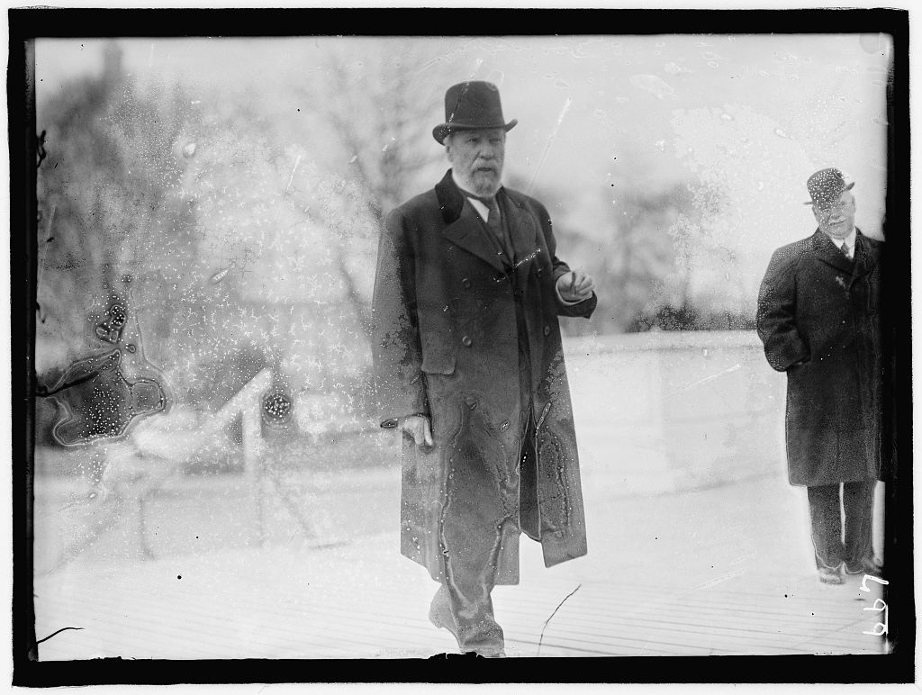 A black and white outdoor photograph of a thickset older man in a suit, long coat, and hat. He has a white beard and mustache.