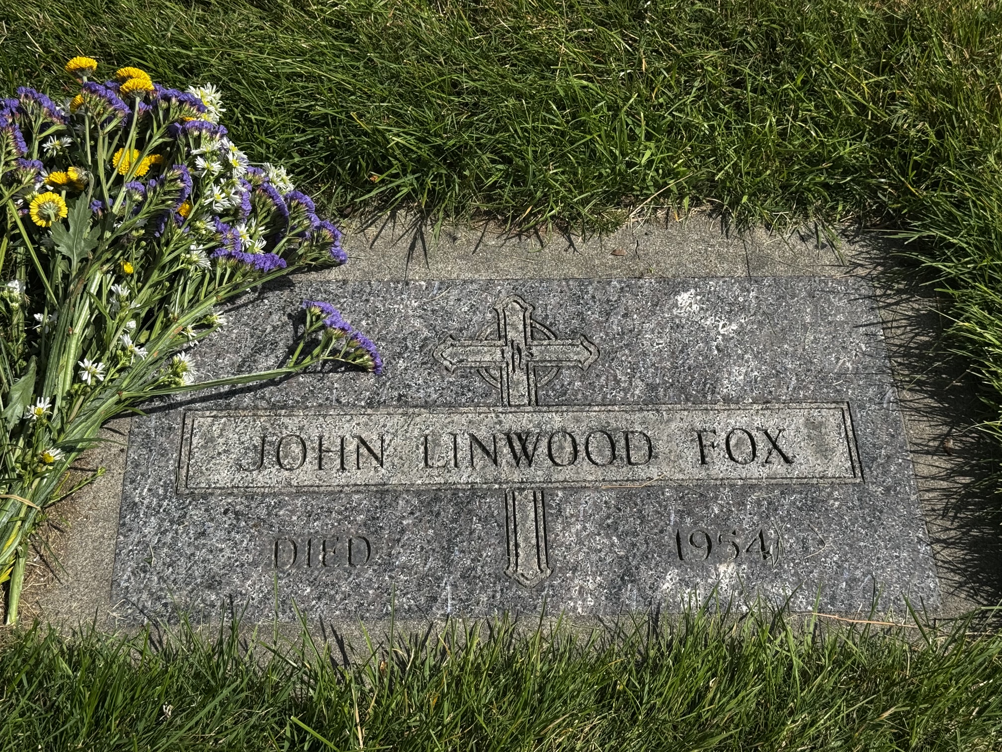 A flat gravestone in the grass with flowers on it. It has a cross with IHS at the top and the name John Linwood Fox. The bottom text reads "died 1954." No birth year is given.