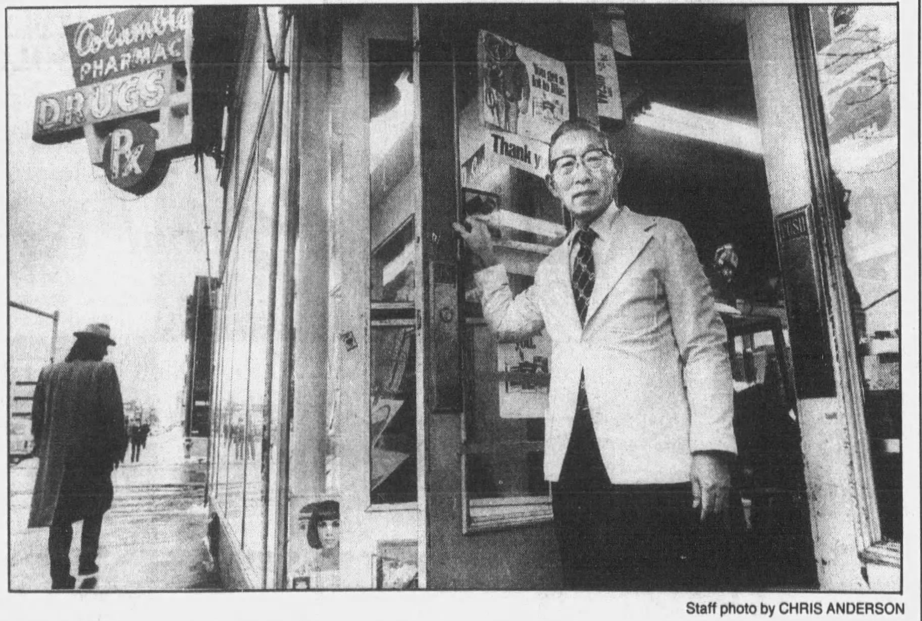 A black and white newspaper photo of an older Japanese man wearing a light-colored coat, a tie, and glasses. He is holding open the door of a building that has "Columbia Pharmacy" on a sign outside.