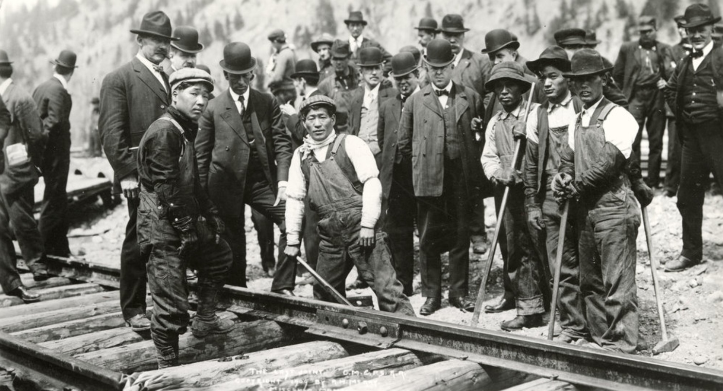 A collection of white men in suits and hats are standing around a railroad track. On the track and in the front of the picture are five Japanese men in work clothes and caps. Some of them hold tools.